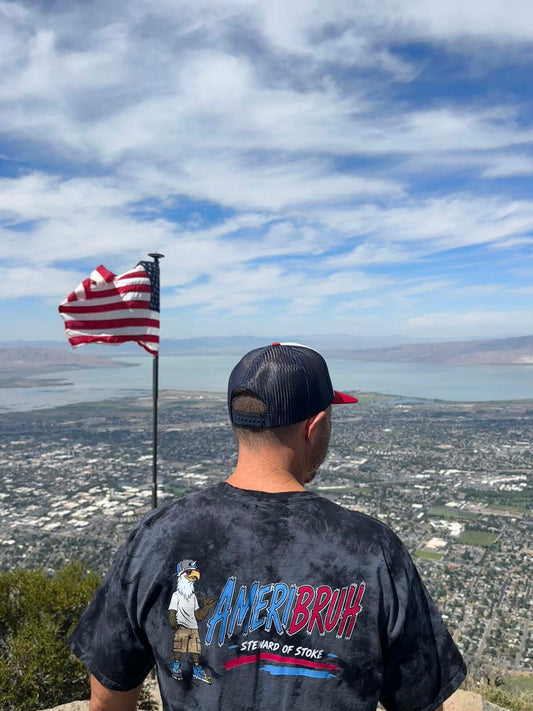 Person wearing Ameribruh black tie dye tee at mountain summit with American flag